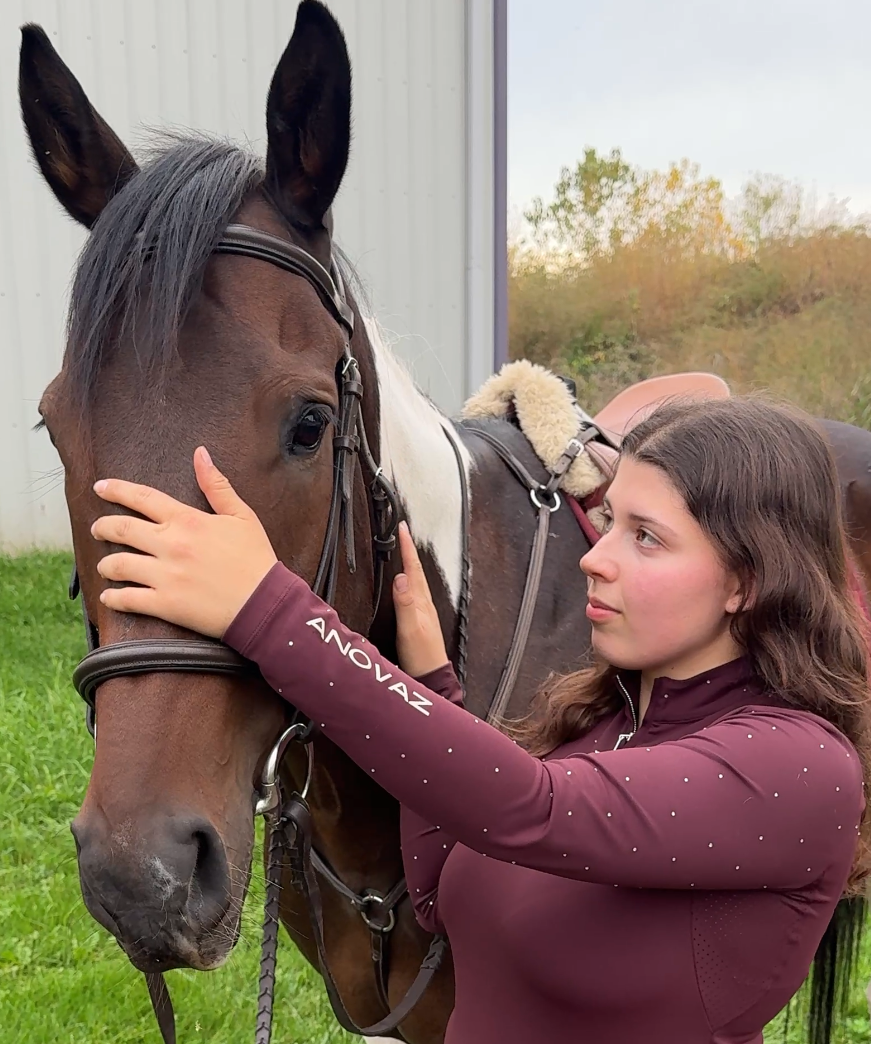 Woman in equestrian attire standing next to a horse on a grassy area.