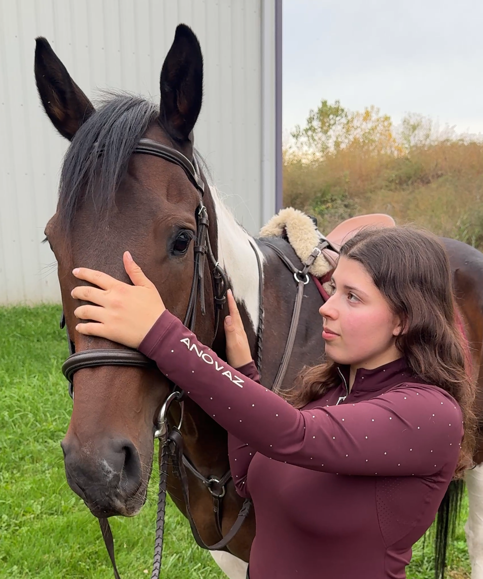 Woman in equestrian attire standing next to a horse on a grassy area.